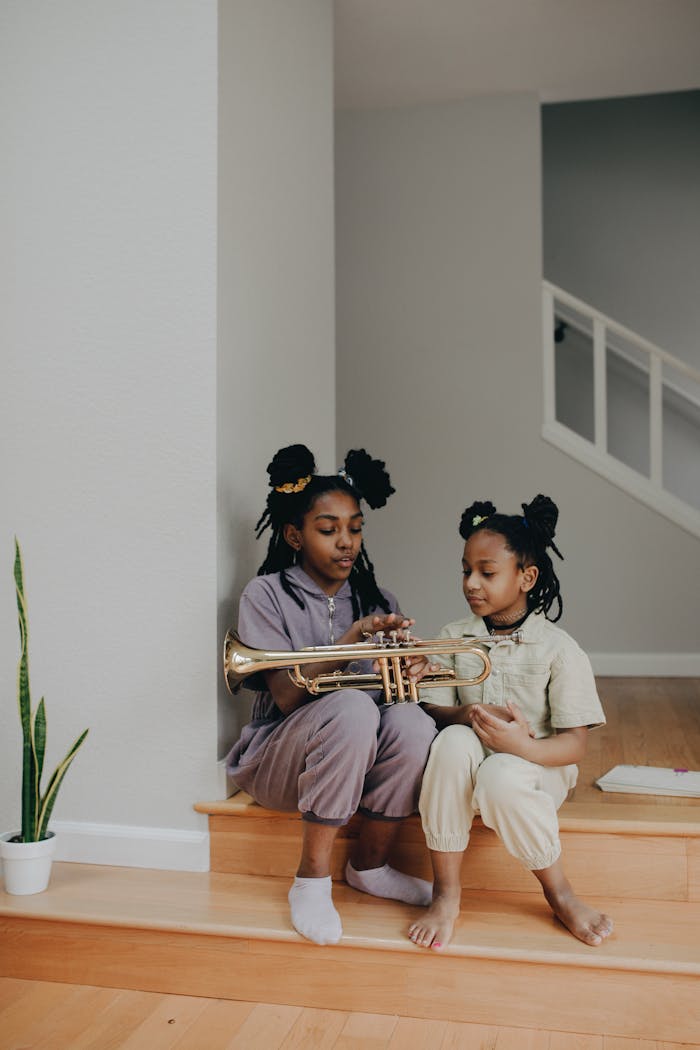 Two sisters sitting indoors, learning to play the trumpet together.